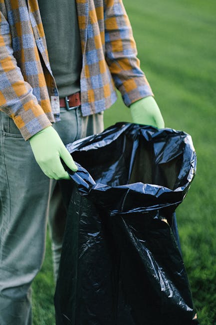 A person standing outdoors on a grassy area, wearing a checkered shirt in shades of yellow and blue over a dark green top, with green rubber gloves and grey trousers, holding a black rubbish bag by it
