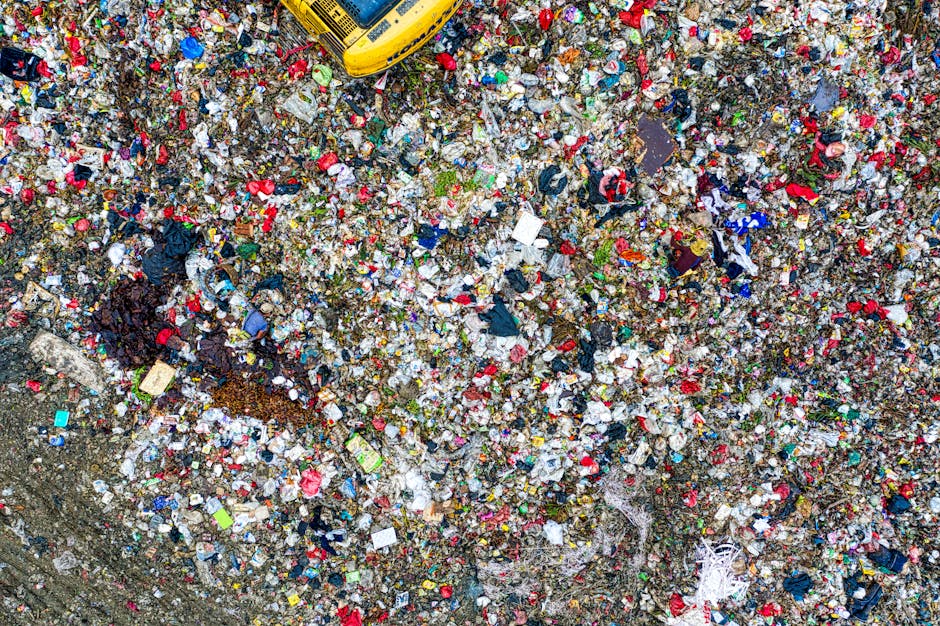 An aerial view of a large landfill site filled with a dense layer of mixed waste, including plastic bottles, packaging, fabrics, paper, and discarded household items. The waste stretches across the en