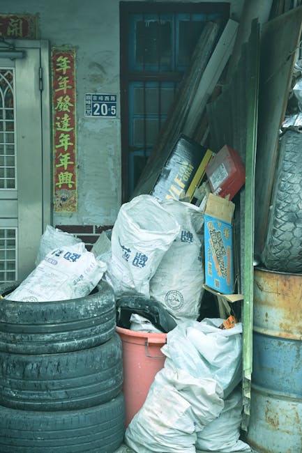 An aerial view of a large landfill site filled with a dense layer of mixed waste, including plastic bottles, packaging, fabrics, paper, and discarded household items. The waste stretches across the en