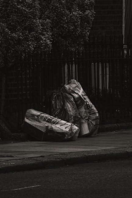 A large pile of mixed rubbish and waste materials, including cardboard boxes, plastic bags, paper, and discarded packaging, is overflowing from a central grey recycling bin labeled 'mixed paper & card