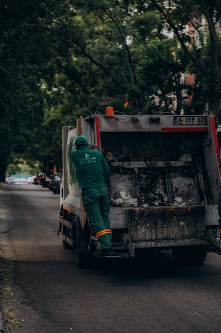 A sanitation worker dressed in green protective overalls, including a helmet and reflective orange stripes on the pant cuffs, is standing on a residential street curbside, loading or inspecting the re