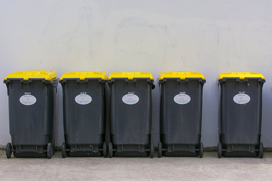 A sanitation worker dressed in green protective overalls, including a helmet and reflective orange stripes on the pant cuffs, is standing on a residential street curbside, loading or inspecting the re